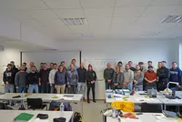 Eine große Gruppe von rund 25 Teilnehmerinnen und Teilnehmern steht in einem Unterrichtsraum zusammen. Vor ihnen stehen Tische mit Lernmaterialien. In der Mitte hängt ein Whiteboard mit der Aufschrift „Industrie Meister“.