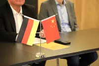 Zwei Tischfähnchen – die deutsche und die chinesische Nationalflagge – stehen nebeneinander auf einem Konferenztisch. Im Hintergrund sitzen zwei Männer im Anzug, teils außerhalb des Fokus. Auf dem Tisch liegen Dokumente und ein Smartphone.