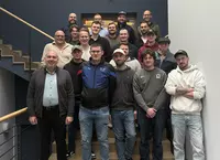 Eine größere Gruppe von Männern, bestehend aus Studierenden und Unternehmensvertretern, steht gestaffelt auf einer Treppe in einem Firmengebäude und posiert für ein gemeinsames Gruppenfoto.