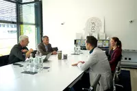 Vier Personen sitzen an einem Konferenztisch mit Getränken und Unterlagen. Zwei Männer auf der linken Seite sprechen mit einem Mann und einer Frau auf der rechten Seite. Im Hintergrund sind Urkunden und ein Wandbild mit dem Schriftzug "BERUF" zu sehen.