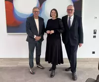 Drei Personen stehen vor einem abstrakten Kunstbild: Dr. Carolin Wagner in der Mitte, flankiert von Vorstand Stephan Koller und Geschäftsbereichsleiter Thomas Skowronek, beim Besuch der Eckert Schulen in Regenstauf.