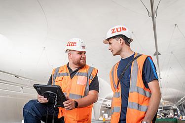 Zwei ZÜBLIN-Mitarbeiter mit Schutzhelmen und Warnwesten im Gespräch auf einer Baustelle, einer von ihnen hält ein Tablet in der Hand.
