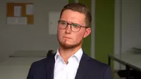 Tim Brücklmayer Junge Mann mit dunkelbonden kurzen Haaren und Brille blickt relativ ernst in die Kamera. Er trägt Bart, eine blaue Jacke und ein weißes Hemd.