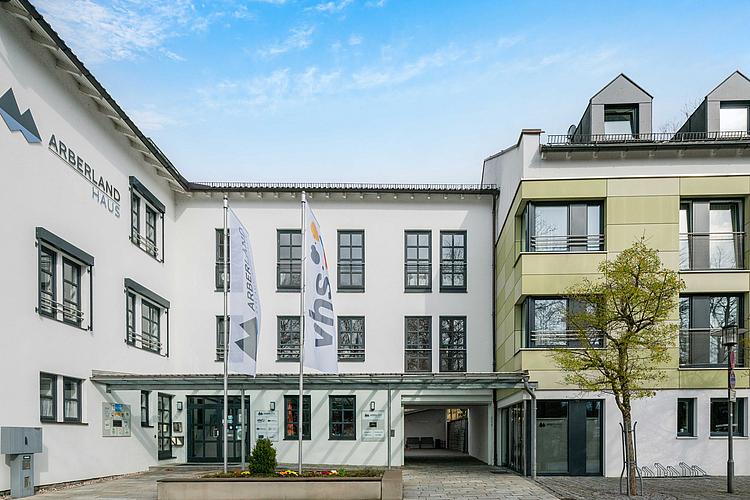 Eingangsbereich des Arberlandhauses in Regen Barrierefreier Eingangsbereich des Arberlandhauses in Regen mit moderner Glasüberdachung, zwei wehenden Fahnenmasten mit den Logos von ARBERLAND und VHS, weißer Gebäudefassade mit schwarzen Fensterrahmen und angebautem grünlichem Flügel auf der rechten Seite. Links am Gebäude ist das ARBERLAND-Logo zu sehen.