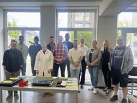 Gruppenbild_AEVO Gruppe von Teilnehmenden steht gemeinsam in einem hellen Raum hinter einem Tisch mit Arbeitsmaterialien; eine Person trägt einen weißen Laborkittel.